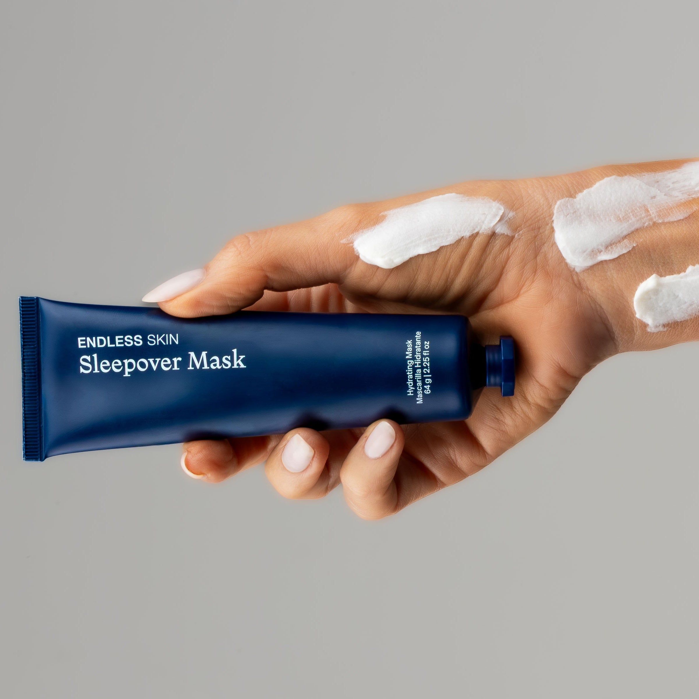 Hand holding a blue tube of 'Sleepover Mask' with white cream on a gray background